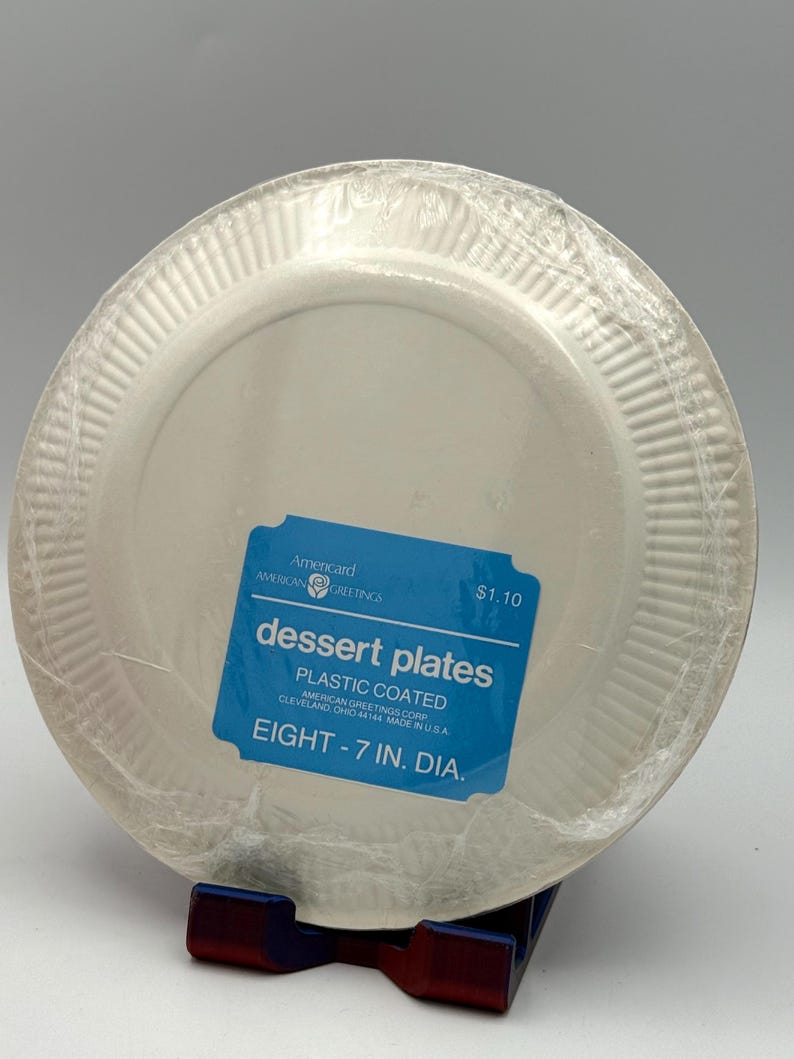 May include: A stack of eight white plastic-coated dessert plates, each 7 inches in diameter, wrapped in clear plastic. A blue label reads "dessert plates" and "plastic coated". The brand is Americard American Greetings.
