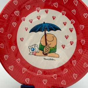 May include: A red plate with a white center featuring a cartoon character under a blue umbrella, surrounded by red hearts. The plate's outer rim is also red with white heart outlines. The plate is supported by a blue and red stand.