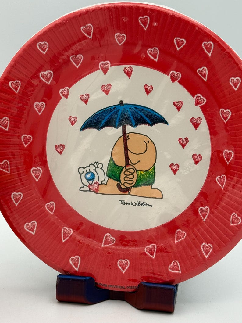 May include: A red plate with a white center featuring a cartoon character under a blue umbrella, surrounded by red hearts. The plate's outer rim is also red with white heart outlines. The plate is supported by a blue and red stand.
