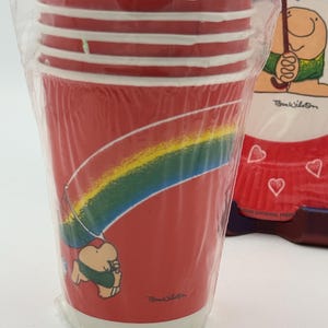 May include: A stack of red paper cups with white stripes and a cartoon rainbow design. The cups are in a clear plastic wrapper. The design includes a cartoon character holding an umbrella. The text "Tom Wilson" is visible.