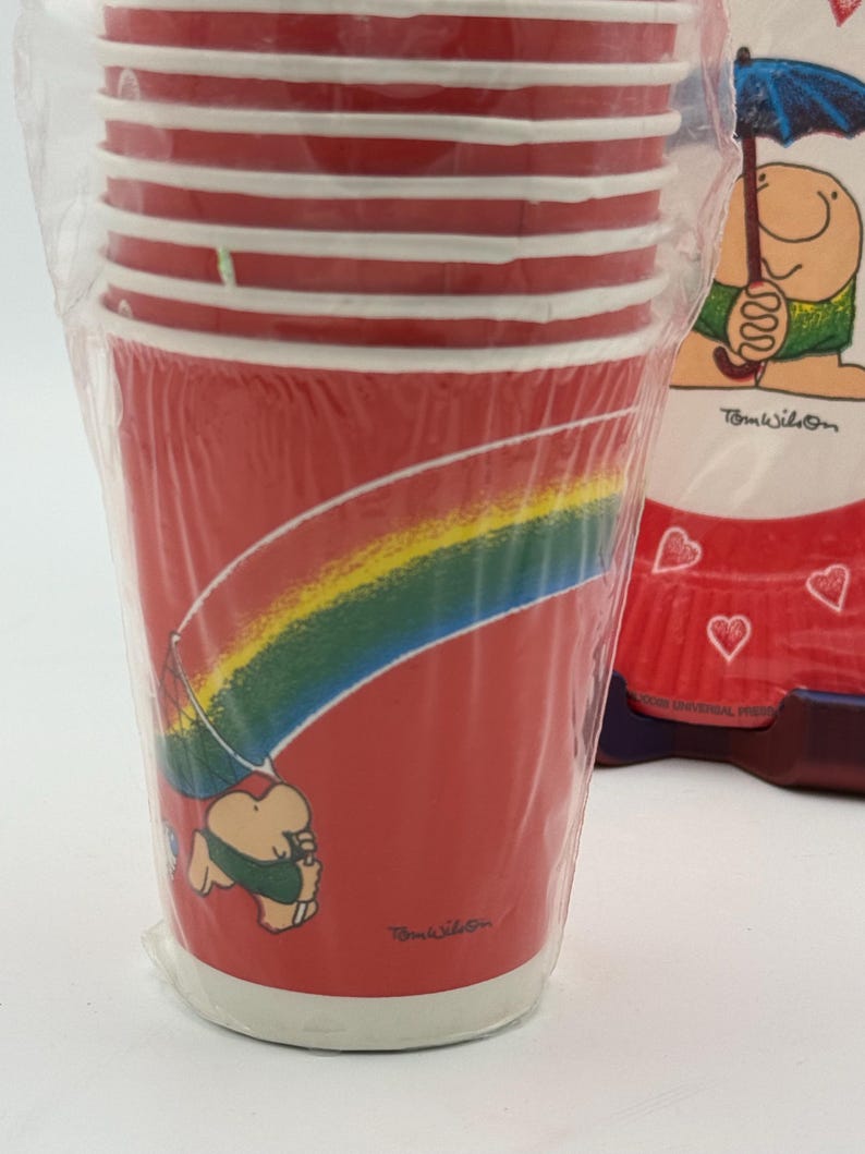 May include: A stack of red paper cups with white stripes and a cartoon rainbow design. The cups are in a clear plastic wrapper. The design includes a cartoon character holding an umbrella. The text "Tom Wilson" is visible.