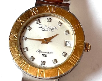 May include: A gold and silver two-tone Bulova quartz watch with a white face and Roman numeral hour markers. The watch has a date window at the 3 o'clock position and the words "Bulova Quartz" and "Superior 2000" printed on the face. The watch has a two-tone band with a gold top and silver bottom.