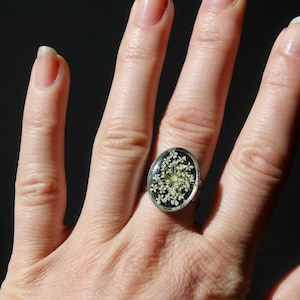 May include: A silver-toned ring with an oval-shaped, resin-filled setting. The setting contains a cluster of small, white flowers against a black background. The ring is worn on a hand with well-manicured nails, set against a dark background.