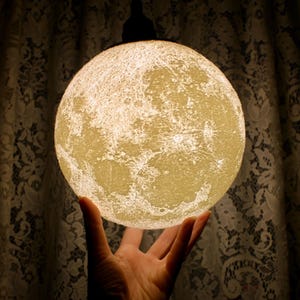May include: A glowing, spherical lamp resembling the moon, held in a hand. The lamp has a textured surface with craters and is illuminated from within. The background features a patterned curtain.