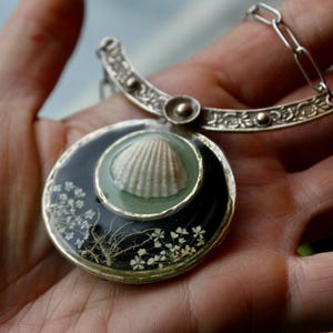 May include: A silver-toned pendant necklace featuring a circular black resin base embedded with small white pressed flowers. A central light green circular inlay holds a small white seashell. The pendant hangs from a decorative silver-toned metal arch.