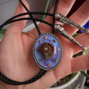 May include: A bolo tie with a black braided cord and silver-toned tips. The oval pendant features a fossilized ammonite set against a blue and purple background, framed by a silver rope-like border. Small, gold-colored accents are scattered around the fossil.