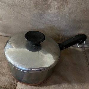 Pode incluir: Uma panela de prata com uma alça e uma tampa pretas. A tampa tem uma pega preta. A panela tem um fundo de cobre.