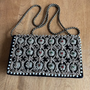 Peut inclure: Une pochette en velours noir avec un motif floral brodé or et argent. Le sac a une bordure perlée et une longue bandoulière noire.