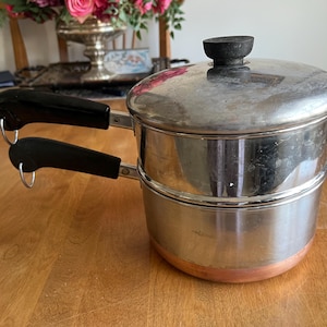 May include: Stainless steel steamer pot with a copper base and black handles. The pot has a lid with a black knob. The steamer pot is on a wooden table.