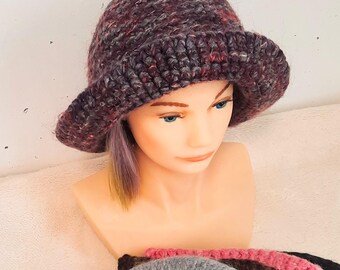May include: A chunky crochet bucket hat in a variegated dark purple and maroon yarn. Below the mannequin head, a collection of similar crochet hats is displayed in a fan arrangement, featuring colors including bright pink, beige, cream, grey, brown, and black.