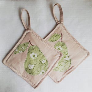 May include: Two square pot holders with a pale pink background. Each has a green pear applique with white floral details and a brown stem. A loop is attached at the top for hanging.