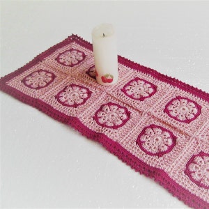 May include: A handmade crochet table runner in shades of pink and magenta. The runner features a repeating floral pattern and a scalloped edge. A white candle with a red apple design sits on the runner.