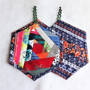 May include: Two hexagon-shaped pot holders, one with a colourful patchwork design and the other with a navy blue floral pattern. Both have green loops for hanging.