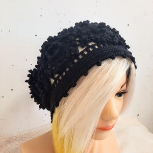 May include: A black crocheted beanie featuring a textured floral pattern with raised knots and a delicate openwork border along the bottom edge. The hat is crafted from dark yarn and displays a complex, layered design with a soft, rounded shape.