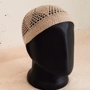 Könnte beinhalten: Eine beige gehäkelte Schädelkappe mit offenem Webmuster. Der Hut wird auf einem schwarzen Schaufensterkopf gezeigt. Die Textur und Farbe des Hutes sind sichtbar, was auf ein handgefertigtes Produkt hindeutet. Die Kappe ist wahrscheinlich für Kopfbedeckungen.