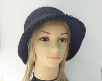 May include: A solid black crochet bucket hat with a textured, looped yarn pattern. The hat features a rounded crown and a downward-sloping brim that encircles the head, providing a consistent dark color and a soft, handcrafted appearance.