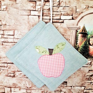 May include: Two pale blue square pot holders featuring a pink gingham apple design with green leaf details. They are suspended by white ribbons. The background is a brick wall.