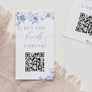 Könnte beinhalten: Zwei weiße Karten mit blauen Blumendesigns und dem Text "KAUFT DER BRAUT EINEN DRINK!" in eleganter Schrift. Jede Karte hat einen schwarzen QR-Code. Die Karten sind wahrscheinlich für eine Hochzeit oder eine Brautveranstaltung.