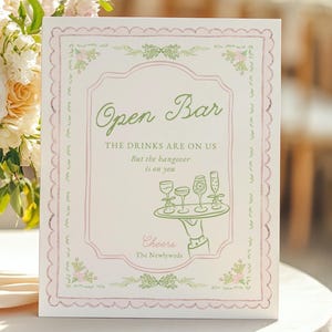 May include: A decorative sign with the text "Open Bar" in green script, framed by a pink border with floral accents. The sign includes the phrase "THE DRINKS ARE ON US But the hangover is on you" and a "Cheers" message.