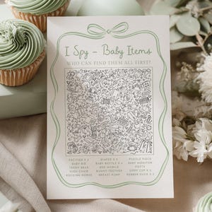Könnte beinhalten: Eine Babyparty-Spielkarte mit dem Text "I Spy - Baby Items" und einer detaillierten Illustration. Die Karte ist von Cupcakes mit grüner Glasur und dekorativen Elementen umgeben. Die Karte enthält eine Liste von zu findenden Gegenständen.