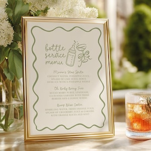 May include: A gold-framed menu with green detailing, listing drink recipes such as "Mama's Glow Spritz" and "Oh Baby Berry Fizz." A glass of iced drink is next to the menu, with white flowers in a vase behind it.