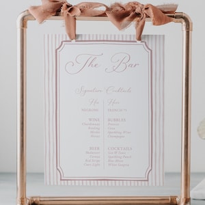 May include: A copper-colored metal bar sign with a menu featuring signature cocktails. The sign has a rectangular frame and is adorned with a decorative ribbon. The menu lists wine, beer, and cocktails. The text "The Bar" is at the top.