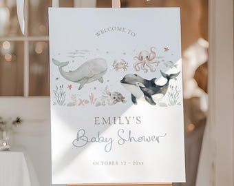 May include: A white sign on a wooden easel, featuring watercolour illustrations of marine creatures, such as whales and an octopus. The sign reads "Welcome to Emily's Baby Shower" with the date below. White balloons are suspended above.
