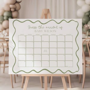 May include: A white sign on a wooden easel with the text "Guess the arrival of BABY WILSON" and a calendar grid. The sign is framed with a wavy green border. A balloon arch in white, beige, and green decorates the background.