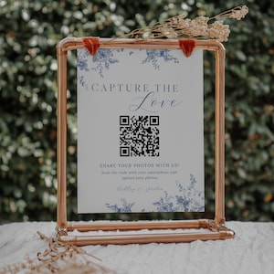 May include: A white sign with blue floral accents and the text "CAPTURE THE LOVE" in black script. A black and white QR code is below the text. The sign is in a copper pipe frame and is sitting on a white surface.