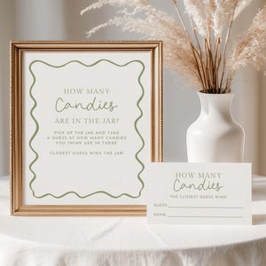 Op de afbeelding: Een ingelijst bord en een kleinere kaart met de tekst "How Many Candies" in een decoratief lettertype. Het bord bevat instructies voor een raadspel. Een witte vaas met gedroogde bloemen staat achter het bord op een wit tafelkleed.