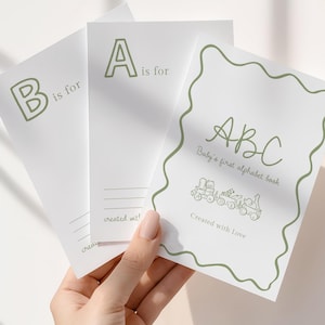 May include: A set of white ABC flashcards with green lettering and illustrations. The cards are held in a hand and feature the letters A and B, along with the text "Baby's first alphabet book" and "Created with Love."