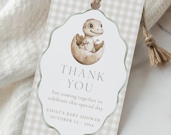 May include: A baby shower gift tag with a watercolor illustration of a dinosaur hatching from an egg. The tag has a gingham pattern and reads "THANK YOU" with event details. A twine string is attached for hanging.