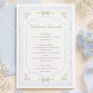 May include: A rectangular wedding weekend details card with a scalloped light blue border and green floral corner accents. The text lists Friday, Saturday, and Sunday event schedules including a rehearsal dinner, ceremony, photos, reception, and brunch.