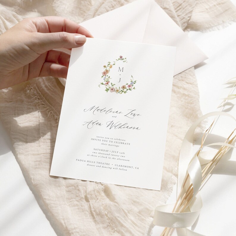 May include: A wedding invitation with a floral wreath design featuring the initials "M" and "J". The invitation announces the marriage of Medeleine Love and Alen Wilkinson on Saturday, July 2nd, 2022 at three o'clock in the afternoon at Padua Hills Theatre in Claremont, CA. Dinner and dancing will follow.