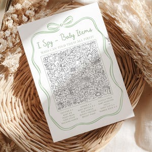 May include: A white card with a green border and text that reads "I Spy - Baby Items". The card features a detailed black and white illustration of baby-related objects. The card is placed on a woven basket.