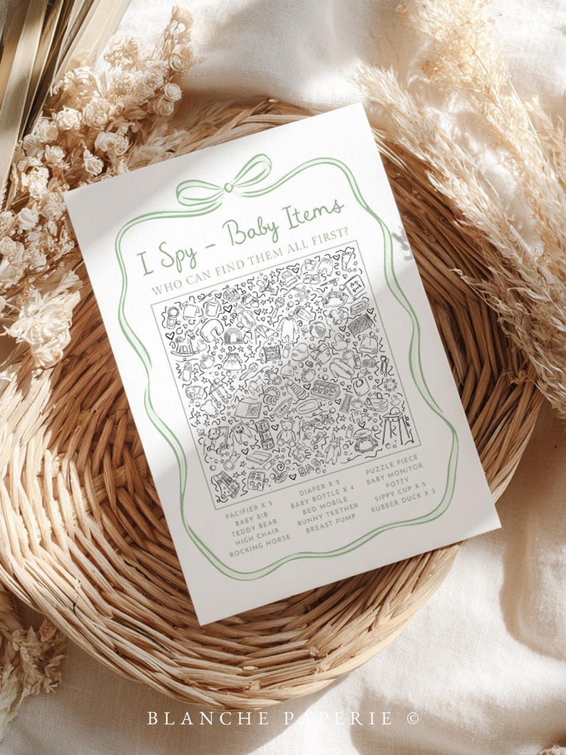 May include: A white card with a green border and text that reads "I Spy - Baby Items". The card features a detailed black and white illustration of baby-related objects. The card is placed on a woven basket.