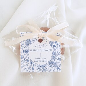 May include: A white favor bag with a blue and white floral design tag tied with a cream ribbon. The tag says "Leigh's Bridal Shower" and "Thank You for Coming!"