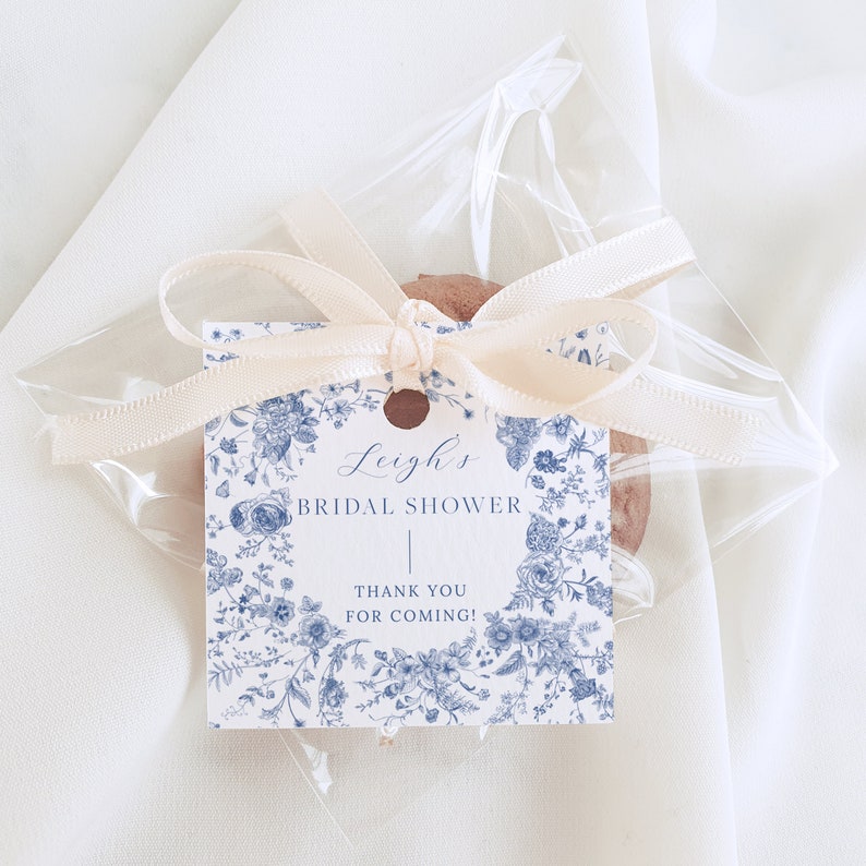 May include: A white favor bag with a blue and white floral design tag tied with a cream ribbon. The tag says "Leigh's Bridal Shower" and "Thank You for Coming!"