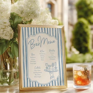 Op de afbeelding: Een ingelijste menukaart met een gouden rand en een blauw-wit gestreepte achtergrond. Het menu bevat bier, wijn en cocktails. Rechts staat een glas amberkleurige drank met ijs en rozemarijn.