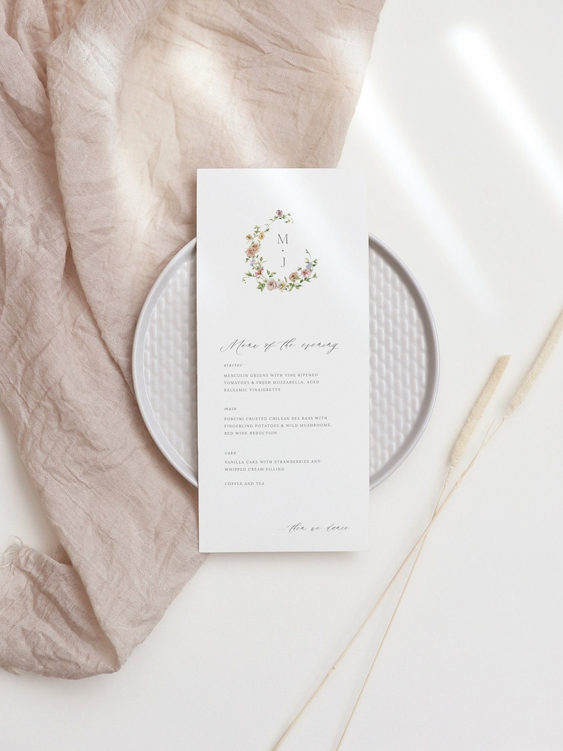 May include: A wedding menu card with a floral wreath design and the initials "M & J" in the center. The menu includes a starter, main course, cake, and coffee and tea. The text "More of the evening" is written in cursive at the top of the card.