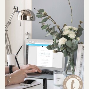 May include: A workspace with a laptop displaying a wedding invitation template. A silver desk lamp, a vase of white flowers and eucalyptus, a candle, and a smartphone are also on the desk. The text "EDIT WITH templett" is visible.