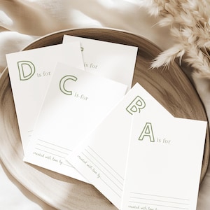May include: A wooden plate holds several white cards with the letters A, B, C, and D in a soft green font. Each card reads "is for" and "created with love by". The cards are arranged on a neutral-toned surface.