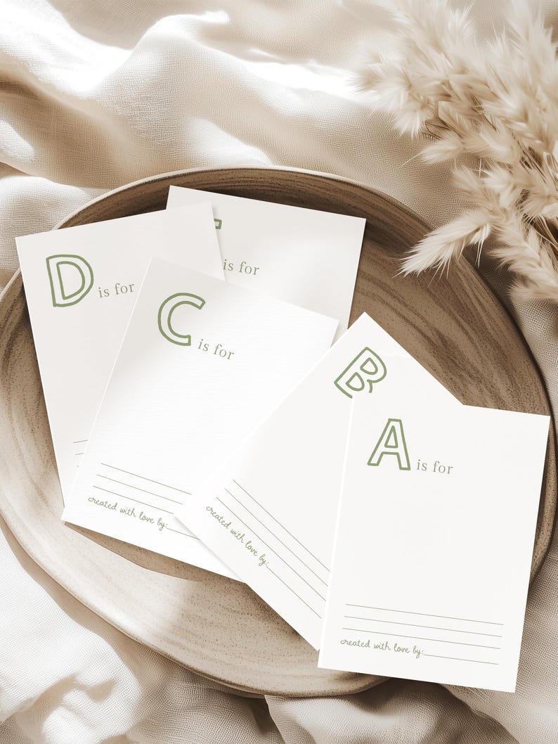May include: A wooden plate holds several white cards with the letters A, B, C, and D in a soft green font. Each card reads "is for" and "created with love by". The cards are arranged on a neutral-toned surface.