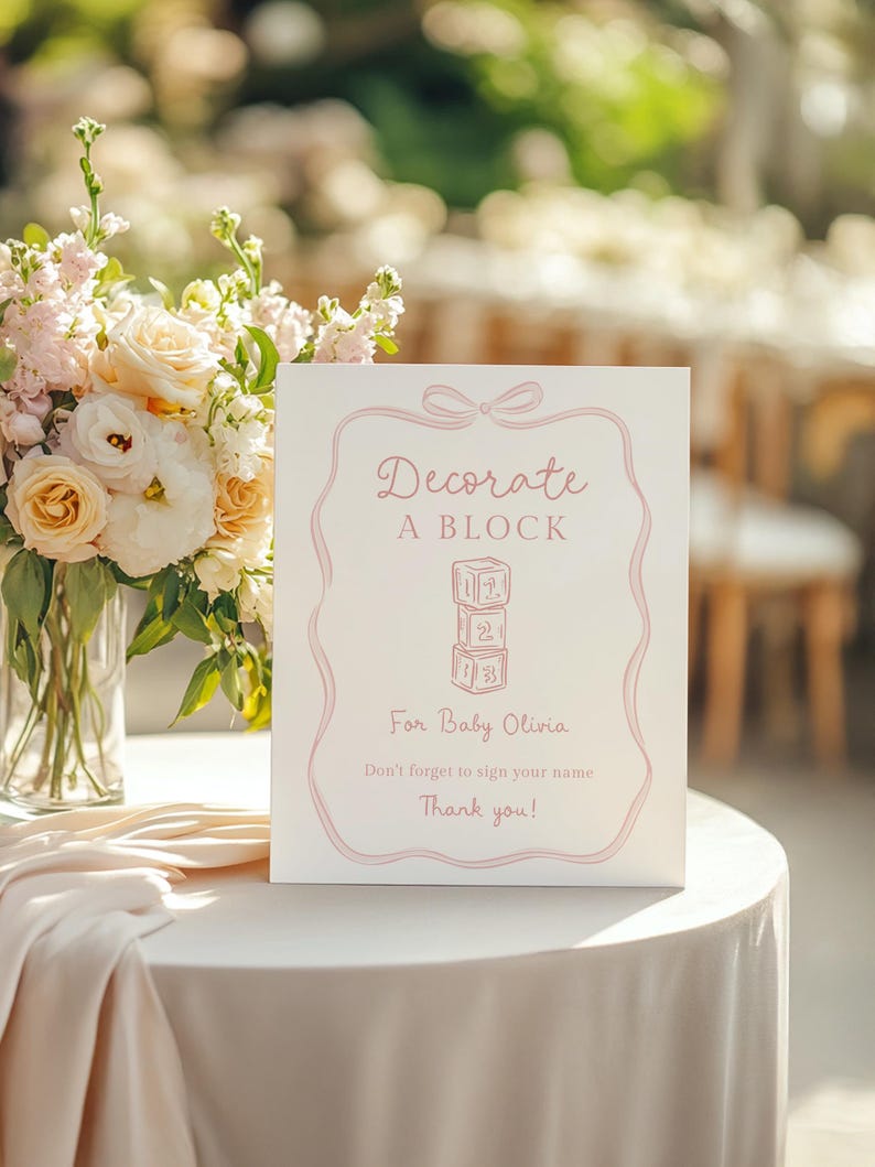 Pu&ograve; includere: Un cartello bianco con testo rosa e bordo rosa che recita "Decorate A BLOCK" con una pila di blocchi giocattolo. Sotto il testo c'&egrave; "For Baby Olivia" e "Non dimenticare di firmare il tuo nome. Grazie!" Il cartello &egrave; su un tavolo bianco.