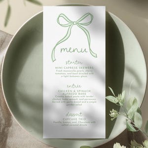 Op de afbeelding: Een menukaart met een groene strik illustratie bovenaan. Het menu bevat voorgerecht-, hoofdgerecht- en dessertopties. Het voorgerecht zijn mini caprese spiesjes. Het hoofdgerecht is kip en spinazie alfredo bake. Het dessert is een cupcake trio.