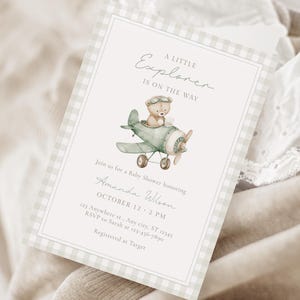 May include: Baby shower invitation with a watercolor illustration of a teddy bear in a vintage airplane. The invitation has a light green gingham border and includes the text "A Little Explorer Is On The Way".