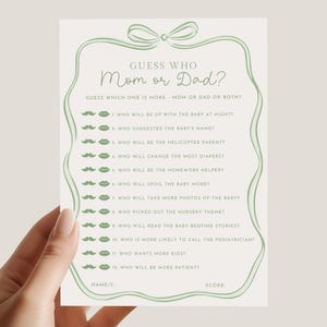 May include: A light green baby shower game card with the text "Guess Who Mom or Dad?" and a list of questions. The card has a decorative bow and border. The card is held by a person.