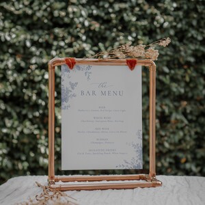 Pu&ograve; includere: Un supporto in tubo di rame con un cartoncino bianco che mostra un menu del bar. Il menu elenca birra, vino bianco, vino rosso, bollicine e bevande firmate. Il cartoncino ha un design floreale con farfalle blu.