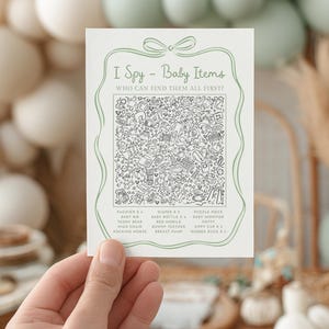 May include: A white card with a green border and text that reads "I Spy - Baby Items". The card features a detailed black and white illustration of baby-related items. The card is held by a hand.