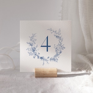 May include: A white card with a blue floral wreath and the number "4" in the centre. The card is on a wooden stand.
